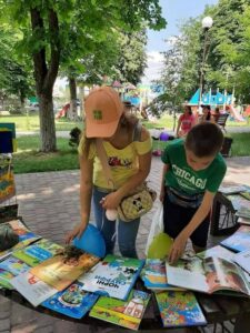 Дівчина в кепці та хлопчик у зеленій футболці розглядають розгорнуті дитячі книжки, що розкладені на лавці у літньому парку. Серед книг видно обкладинку коміксу про Смурфів