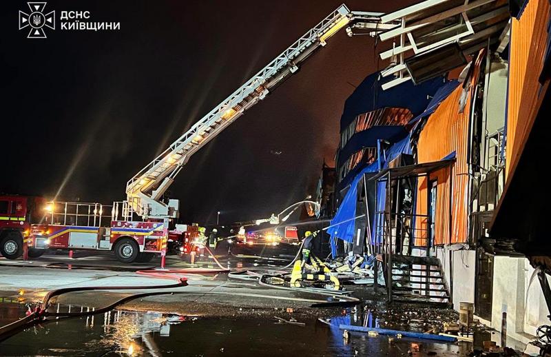 Знімок зроблено вночі. Величезне логістичне приміщення з синьо-жовтим фасадом сильно пошкоджене, видно наслідки влучання та вогонь всередині. Піднята довга пожежна драбина, з якої, ймовірно, подавали воду. На землі працюють рятувальники, розгорнуті пожежні рукави, асфальт мокрий. Працює освітлення спецтехніки. У лівому верхньому куті – логотип "ДСНС КИЇВЩИНИ".