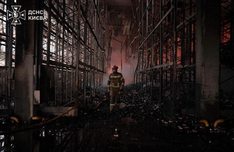Вид зсередини величезного, вщент вигорілого складського приміщення. З обох боків височіють обвуглені металеві стелажі, дах обвалився. По центру, через купи згорілого сміття та калюжі, йде один рятувальник у повному екіпіруванні. На задньому плані, крізь отвори в стінах, видно заграву пожежі. У лівому верхньому куті – логотип "ДСНС КИЄВА".