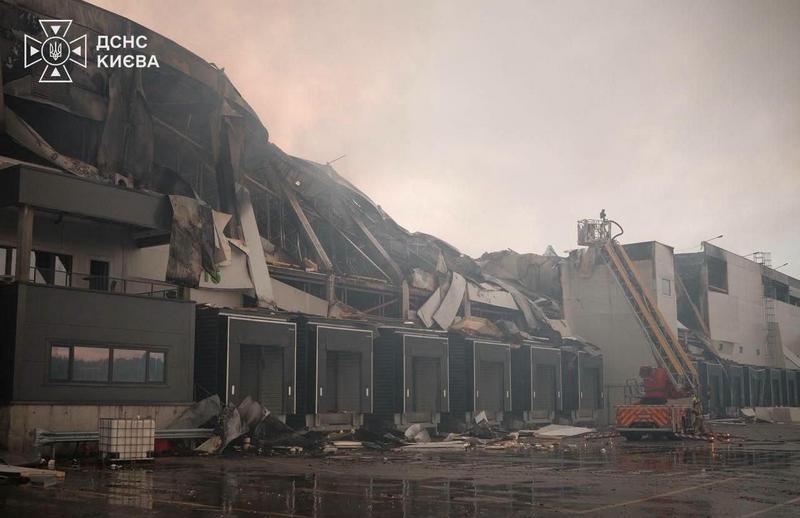 На фото показано значні руйнування великого складського комплексу на Київщині після обстрілу. Його дах повністю провалений, стіни пошкоджені, металеві конструкції зігнуті. Поруч стоїть пожежна драбина ДСНС, рятувальники працюють на висоті. Територія завалена уламками, на землі калюжі. У лівому верхньому куті – логотип "ДСНС КИЄВА".