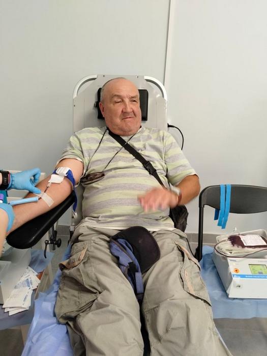Чоловік у смугастій футболці сидить у спеціальному кріслі. Медичний працівник у блакитних рукавичках фіксує катетер на його руці для забору крові.