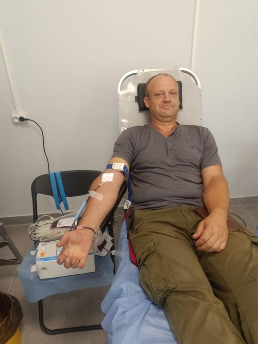 Донор у сірій футболці та штанах кольору хакі сидить у кріслі під час процедури забірного процесу. Поруч на стільці розміщено мішок із зібраною кров'ю.