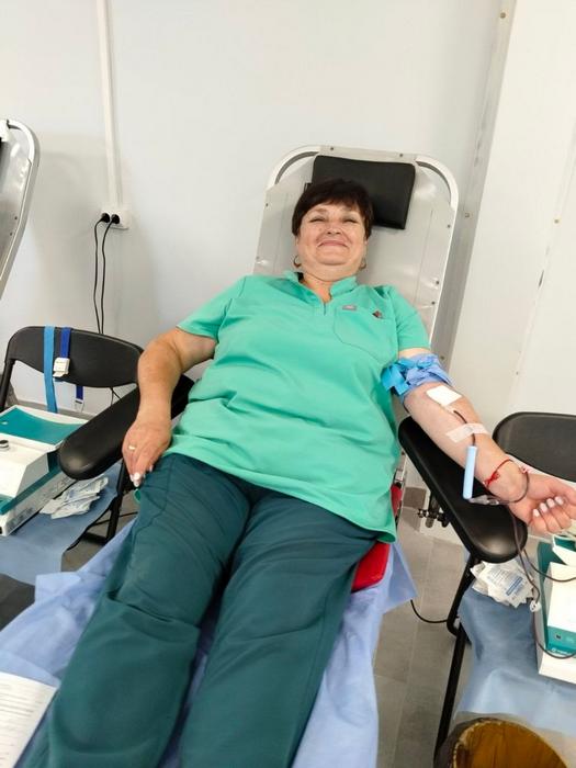 Донорка в салатовій футболці та зелених штанах відпочиває в медичному кріслі під час здачі крові. Рука зафіксована для процедури, поруч знаходиться медична документація.