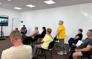 Учасники обговорення різного віку сидять у кабінеті зі світлими стінами та уважно слухають представників селищної ради та консультантів проєкту HOPE.
