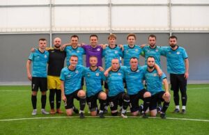 Команда «ФСК Макарів-Saints» позує після перемоги у фіналі: гравці вишикувалися у два ряди (стоячи та на колінах) на зеленому штучному газоні всередині критого манежу.
