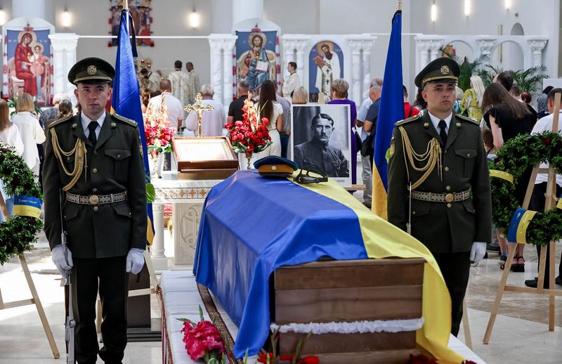 Церемонія прощання з Євгеном Коновальцем у Патріаршому соборі Воскресіння Христового в Києві. У центрі — труна, покрита синьо-жовтим прапором України. Біля труни стоїть почесна варта з двох військових у парадній формі із гвинтівками. Позаду розміщено портрет Євгена Коновальця та державні прапори. На передньому плані — живі квіти.
