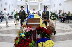 Момент вшанування пам'яті Євгена Коновальця. Чоловік у цивільному одязі став на одне коліно перед труною, що покрита синьо-жовтим прапором, і торкається її руки. Біля труни стоїть почесна варта з двох військових. На передньому плані розміщено багато букетів квітів у синьо-жовтих та червоних кольорах.