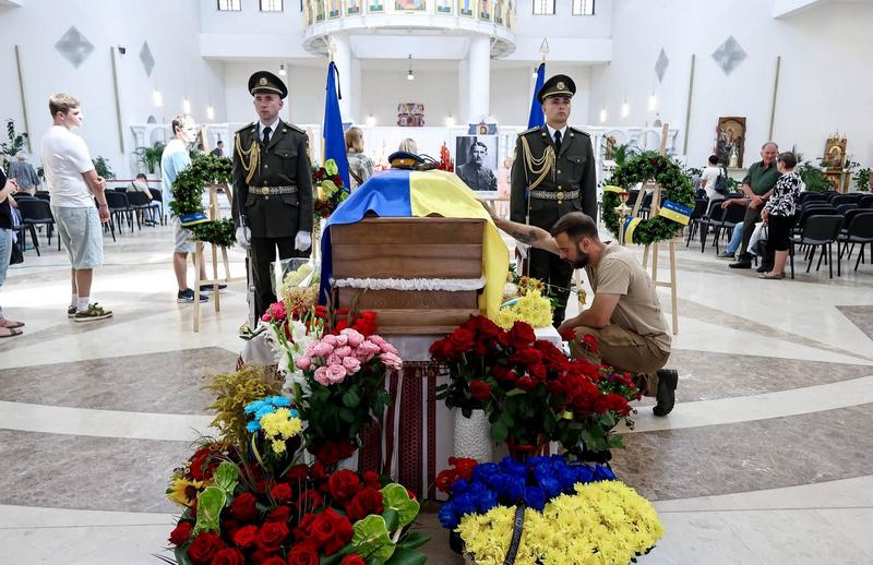 Момент вшанування пам'яті Євгена Коновальця. Чоловік у цивільному одязі став на одне коліно перед труною, що покрита синьо-жовтим прапором, і торкається її руки. Біля труни стоїть почесна варта з двох військових. На передньому плані розміщено багато букетів квітів у синьо-жовтих та червоних кольорах.