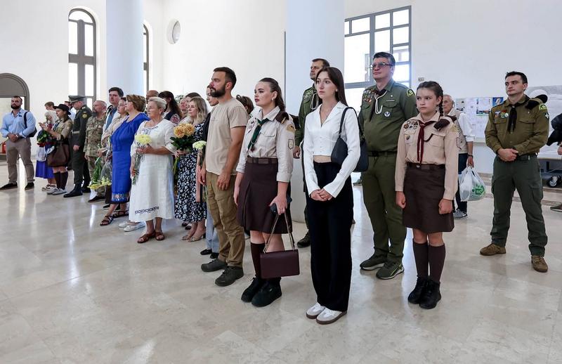 Люди, які прийшли вшанувати пам'ять Євгена Коновальця в соборі. Серед присутніх — пластуни в одностроях (дівчата у світло-коричневих сорочках та спідницях, хлопці в оливкових сорочках), жінки у вишитих сукнях з букетами квітів та інші цивільні та військові особи. Вони стоять у ряду, спостерігаючи за церемонією.