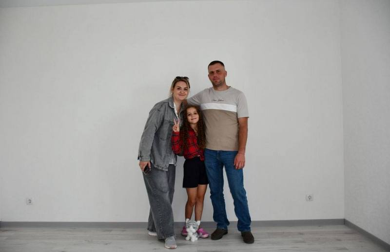 Щаслива родина з трьох осіб — батько, мати та донька — стоять у своїй відновленій квартирі в Макарові. Батько обіймає доньку, а мати посміхається. Кімната готова до заселення, з білими стінами та світлою підлогою.