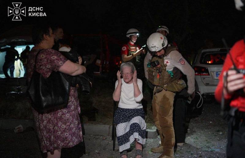 Літня жінка тримається за голову від розпачу, поруч із нею медики та рятувальники на тлі наслідків нічної ворожої атаки.