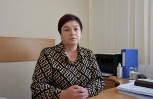 Портрет Валентини Курач, жінки середнього віку з короткою стрижкою, одягненої в блузку з геометричним візерунком. Вона сидить за робочим столом, склавши руки перед собою, на тлі офісного інтер'єру з папками та технікою.