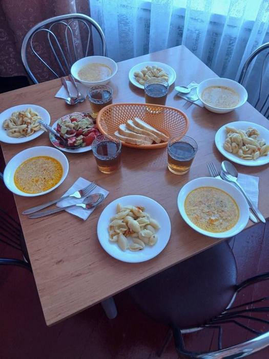 Накритий стіл у шкільній їдальні з тарілками супу, макаронами, овочевим салатом, хлібом та напоями.